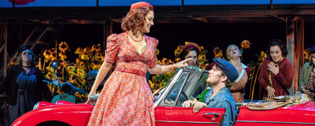 Adina in a red dress beside a vintage red car with Nemorino in The Elixir of Love